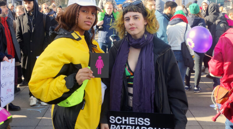 2 people at the International Women's Day in Berlin 2019 with the CO-WC sign and a poster with the inscription "Scheiß Patriarchat Elendiges."