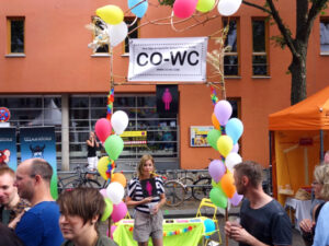 Person in front of CO-WC info booth, with the sign in hand and observed by visitors of the Lesbian and Gay City Festival.